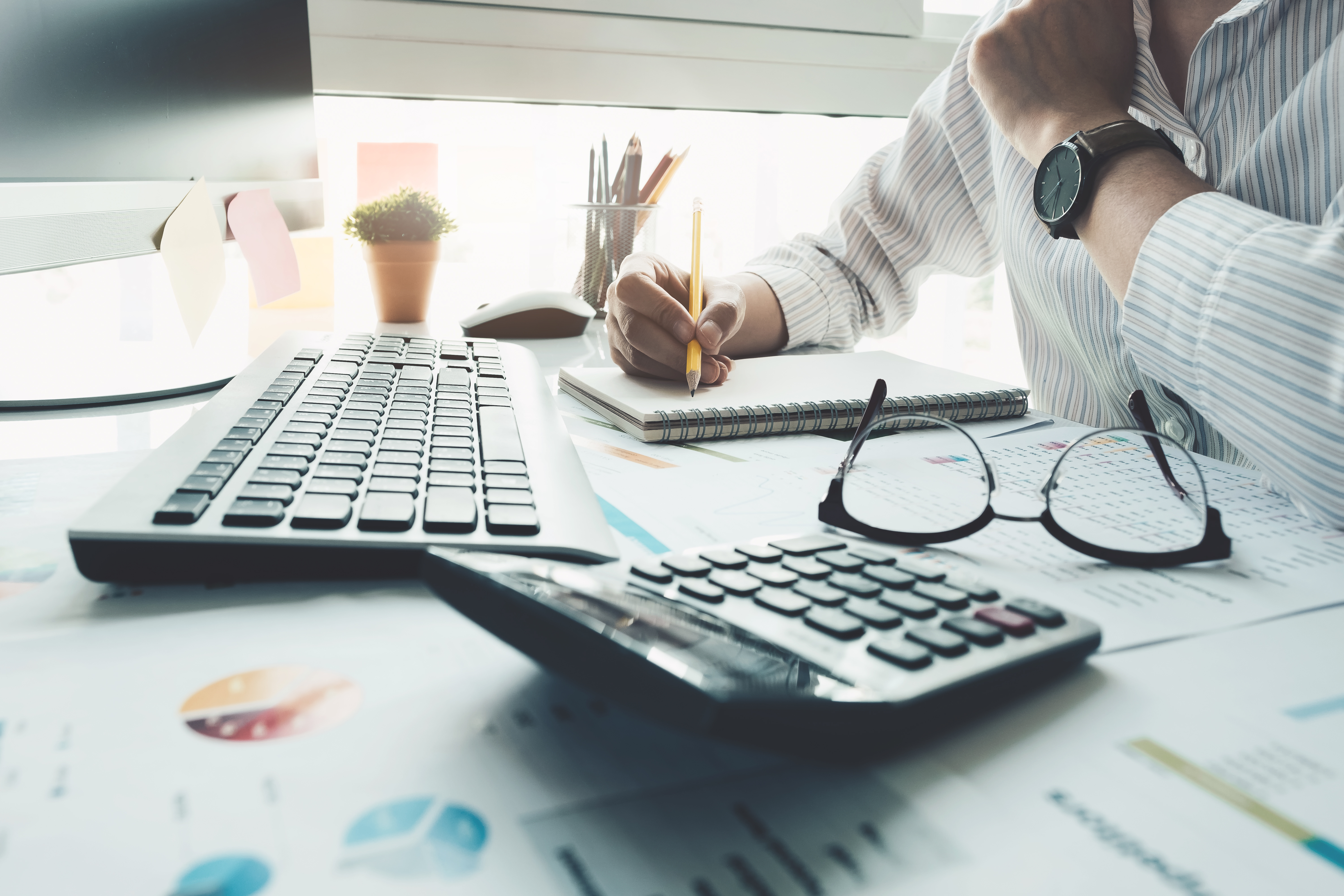  Person writing in a notebook at a desk with a keyboard, calculator, glasses, and financial charts, conveying a work or financial planning setup.