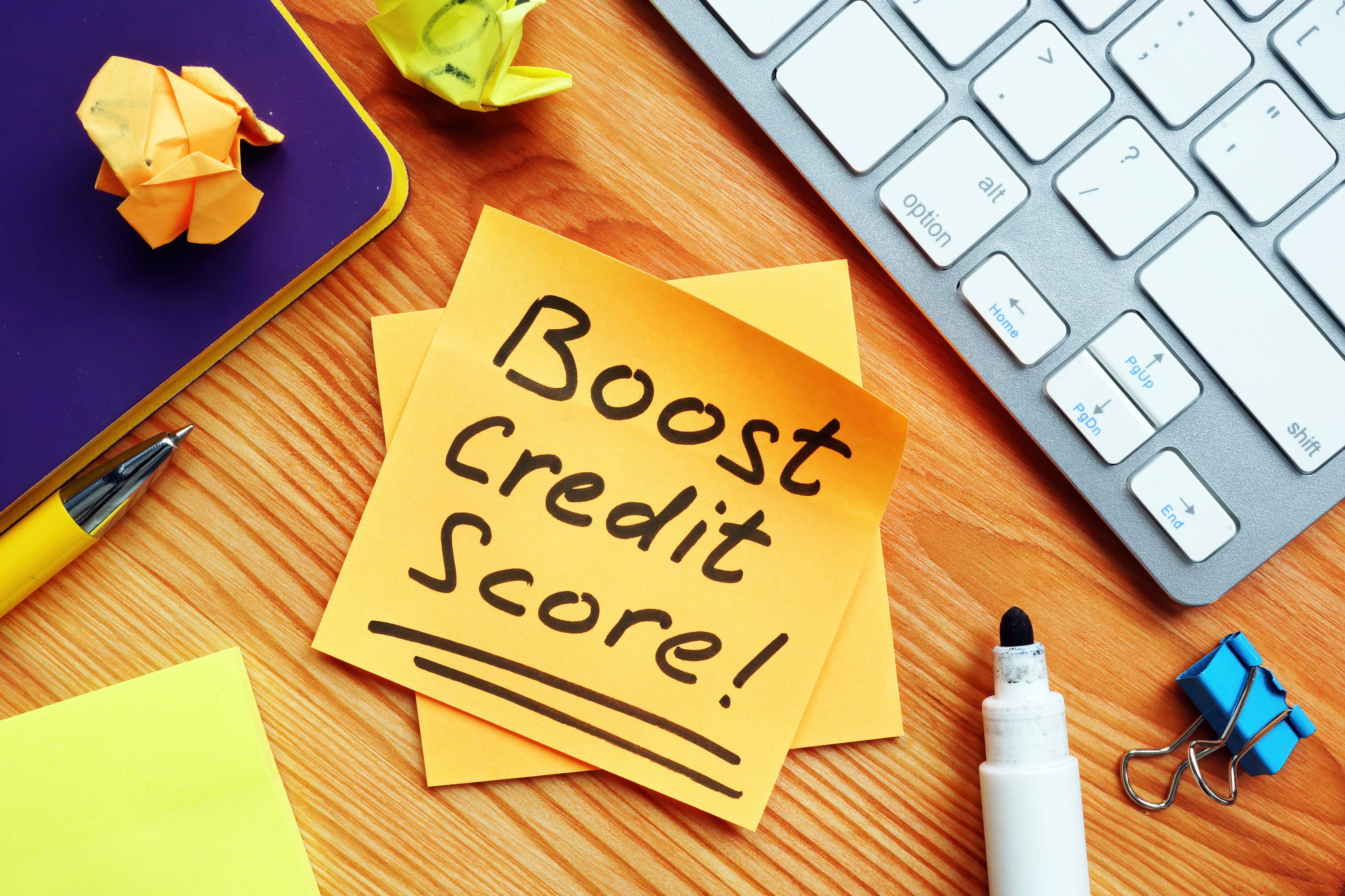 Sticky note with the text 'Boost Credit Score!' on a desk surrounded by a keyboard, pen, and office supplies, symbolizing strategies to improve financial health.