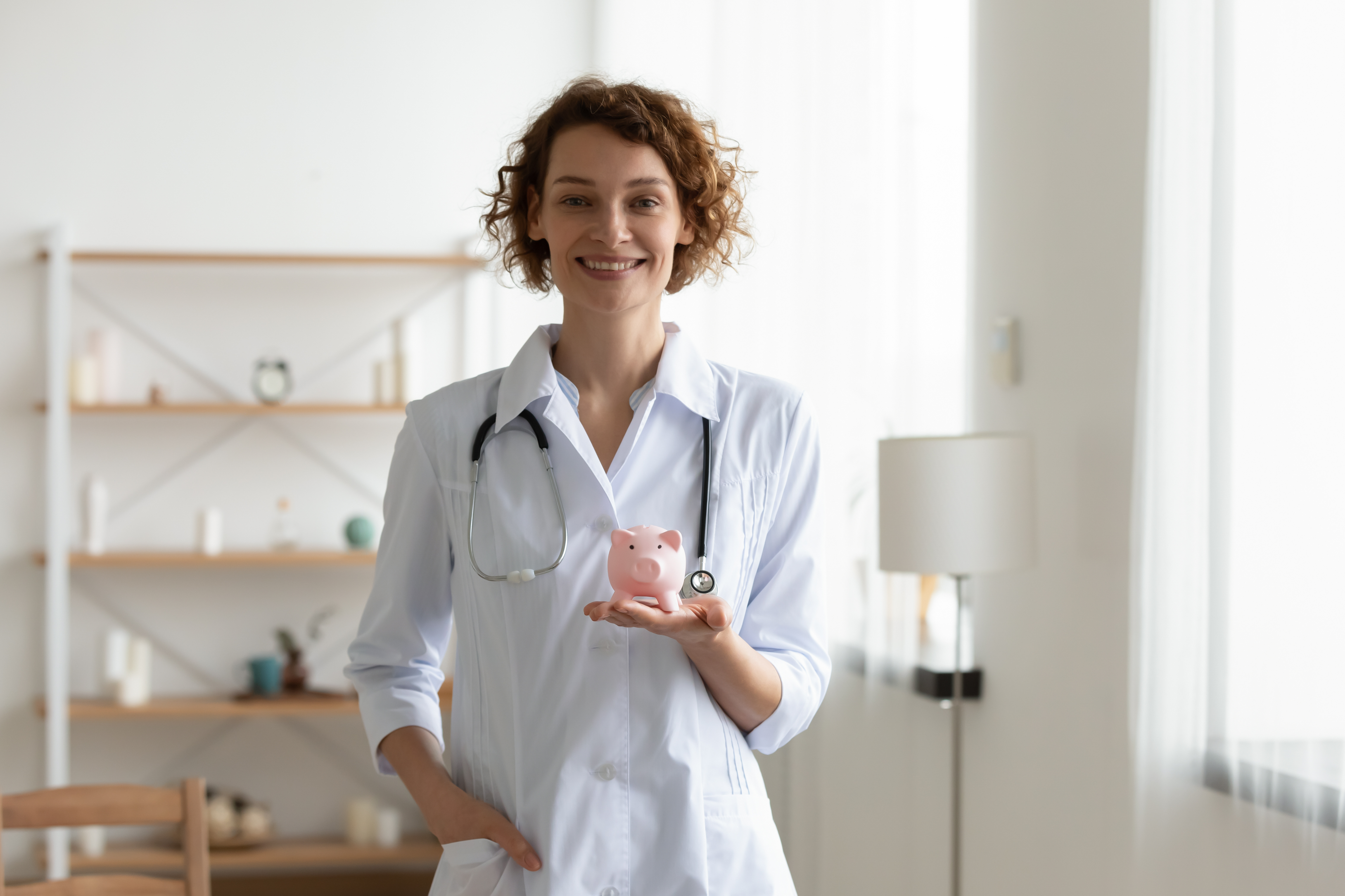 A smiling female doctor wearing a white coat and stethoscope, holding a pink piggy bank, standing in a bright room with shelves and a lamp in the background.