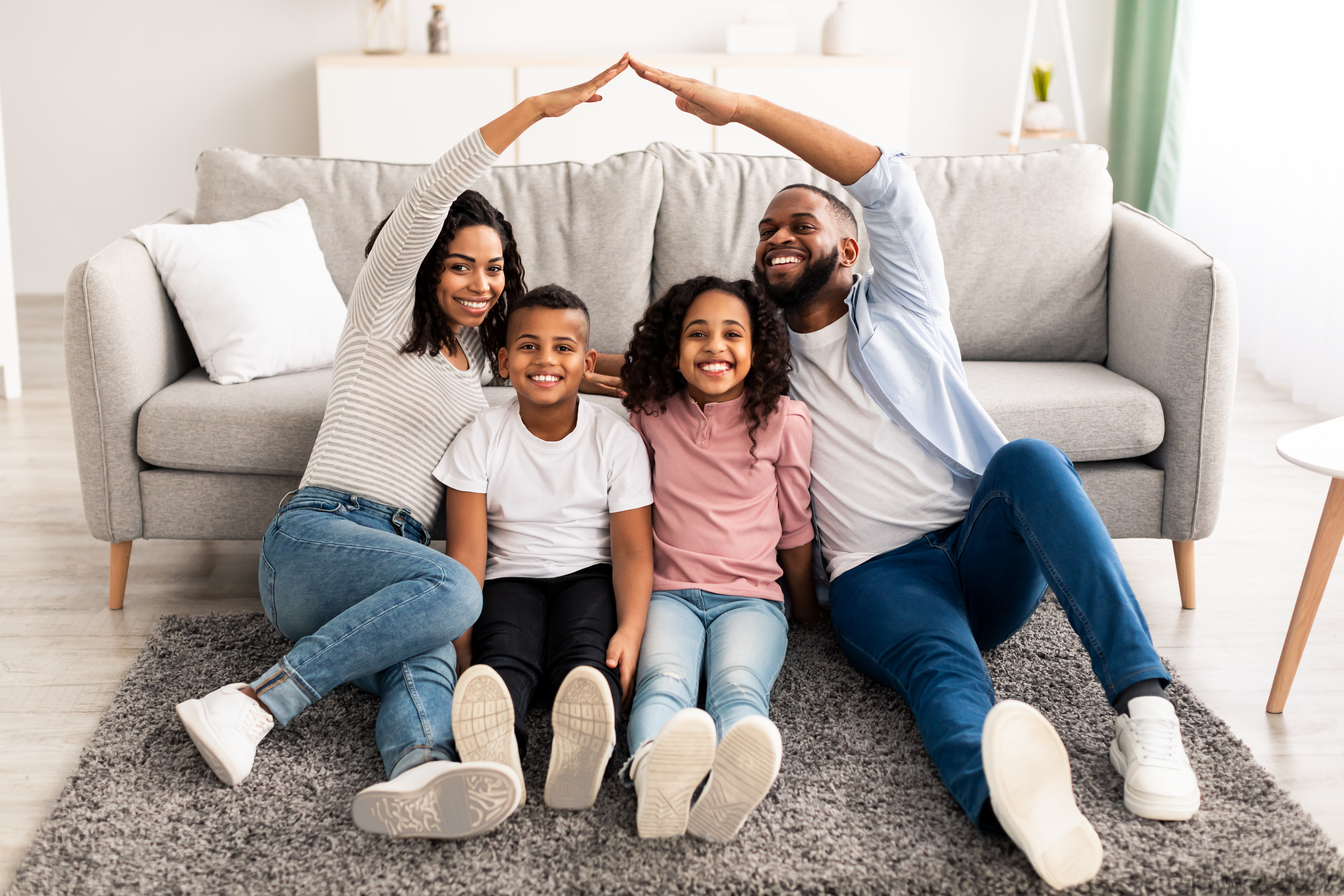 A joyful family of four sitting on a carpet in their living room, with the parents forming a roof-like shape over their two children using their arms, symbolizing love and protection.