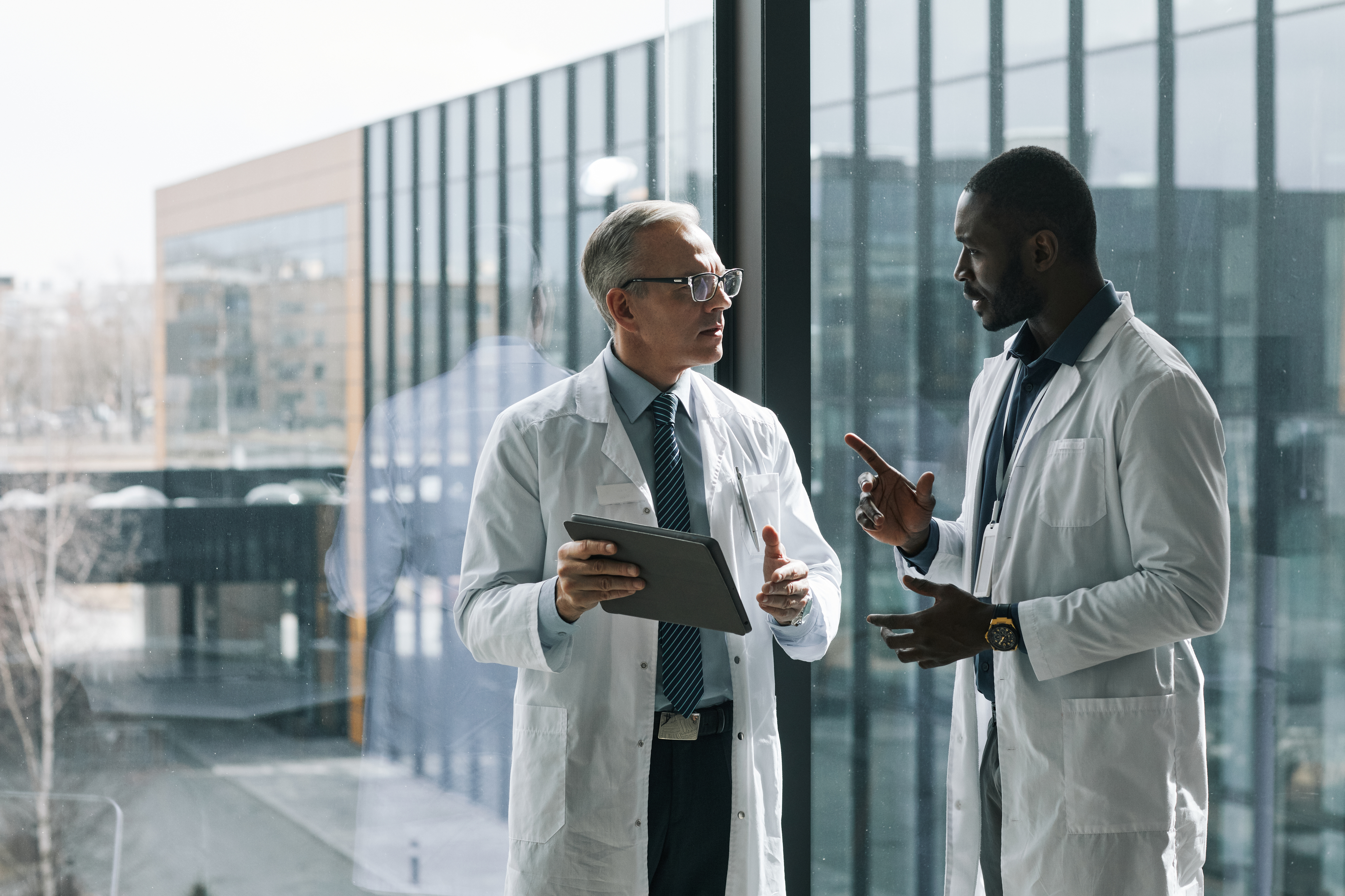 Two doctors in white lab coats standing by a large window in a modern hospital building, engaged in a professional discussion. One doctor holds a tablet, and they appear to be exchanging ideas or consulting on a case.