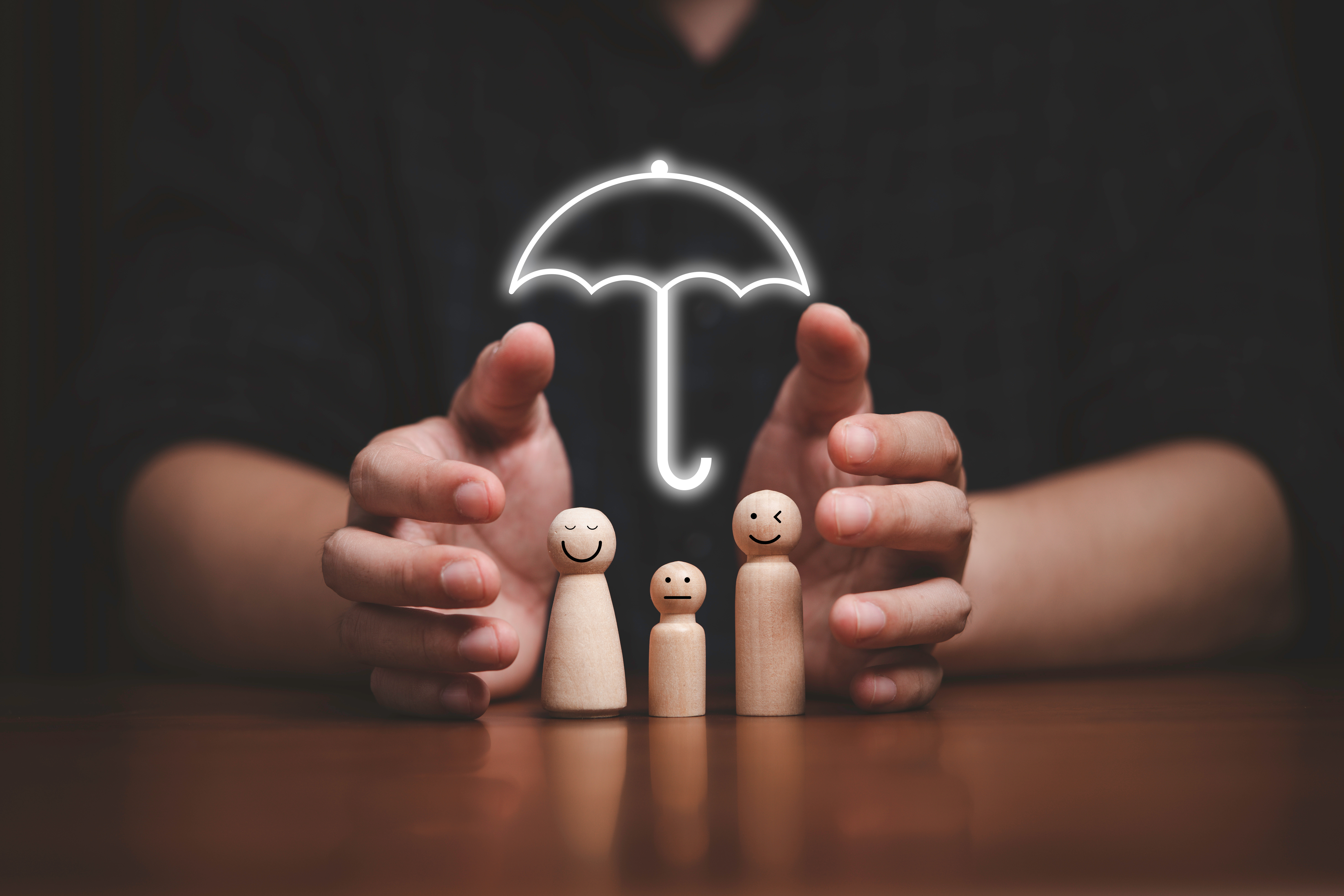 A close-up of hands protecting three wooden figurines with smiley faces under a glowing umbrella icon, symbolizing Life insurance for doctors, care, protection, and safety.