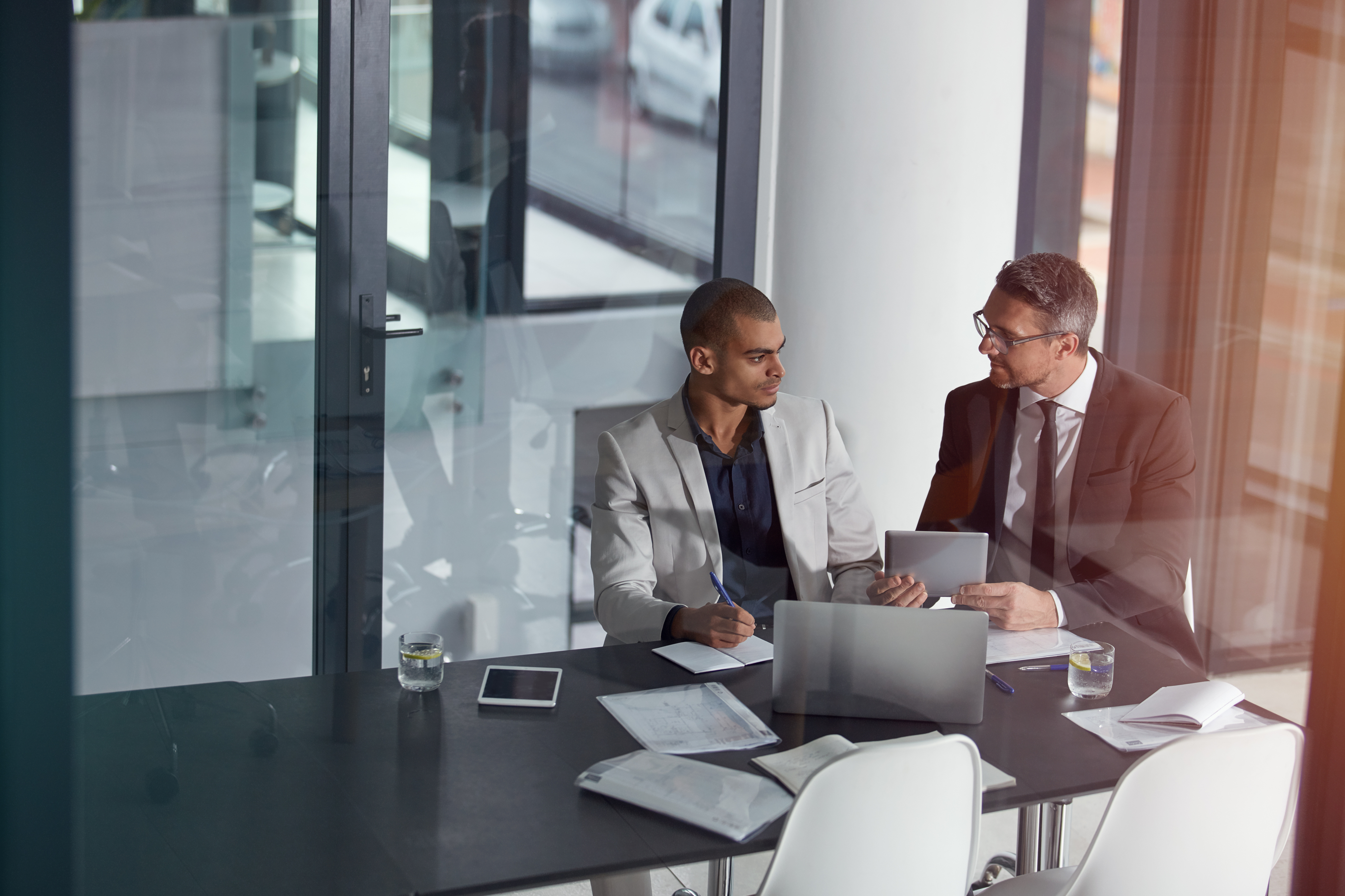 Two men sitting at a table talking over documents with an open laptop. One of them is taking notes, the other is explaining something to him. There is also a glass of water with lemon and a tablet on the table.