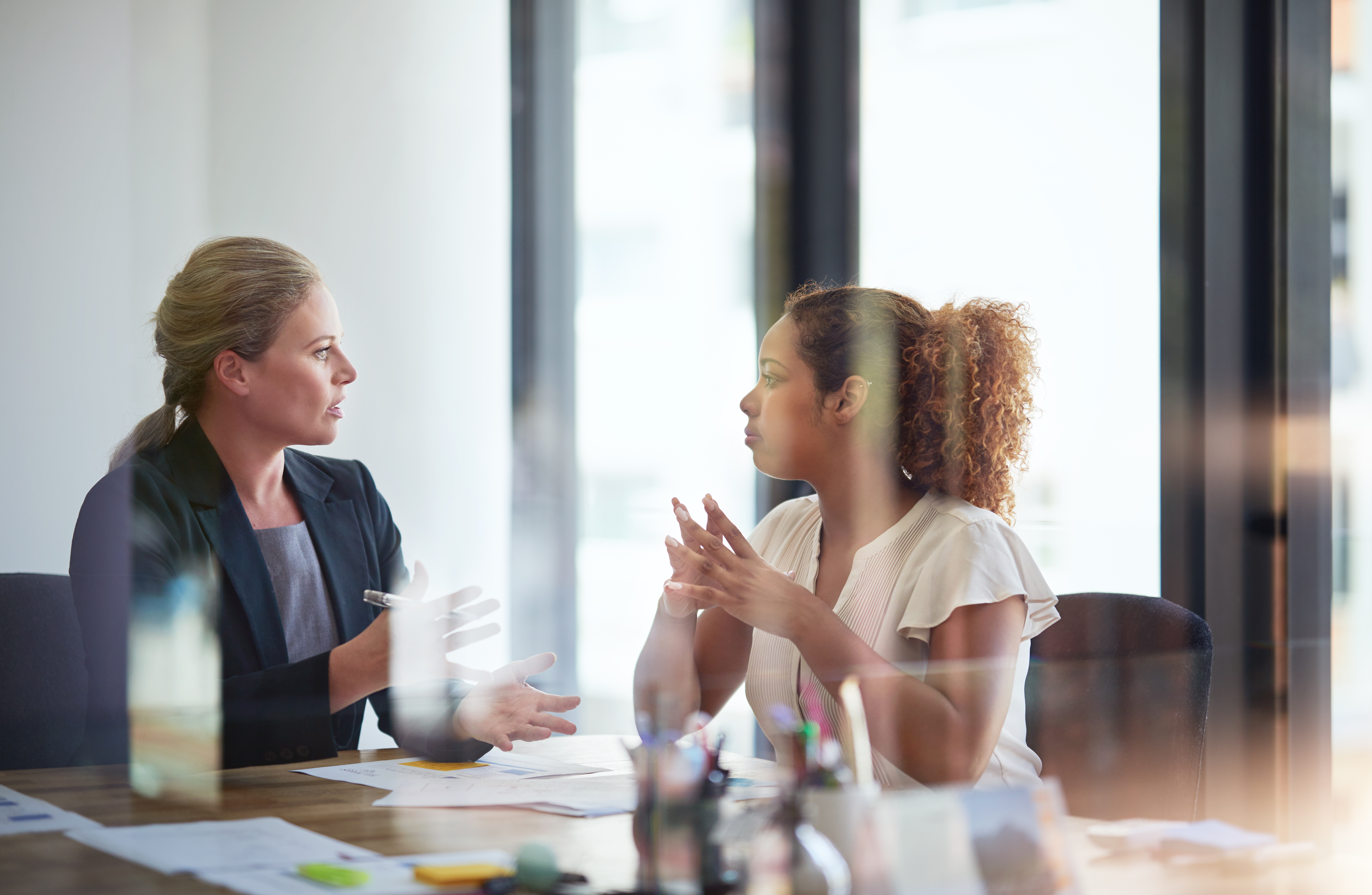  Two businesswomen having a focused discussion in an office setting, with documents on the table, as seen through a glass partition. Building Financial Habi