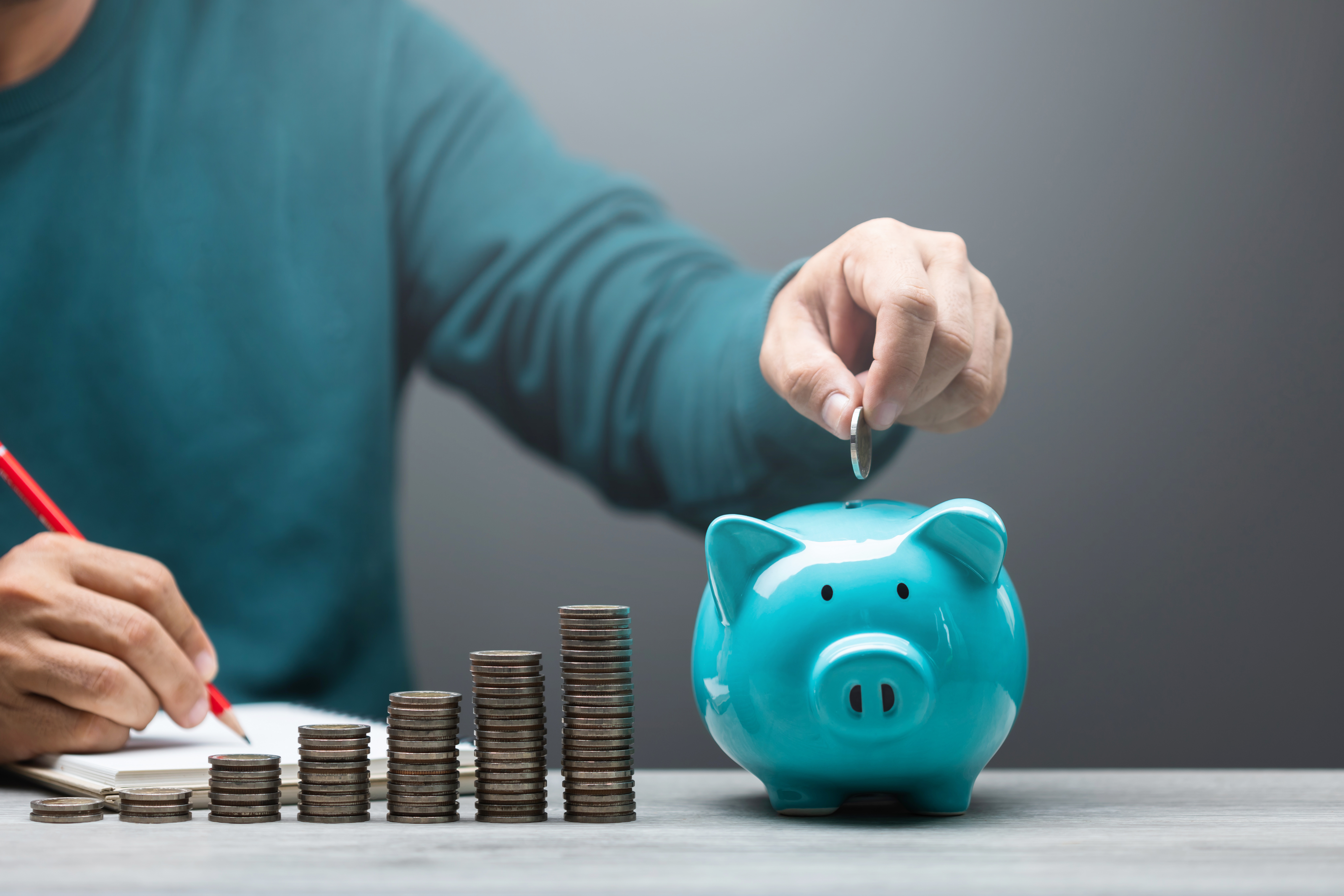 Person dropping a coin into a blue piggy bank with stacks of coins increasing in height, symbolizing savings and financial growth.