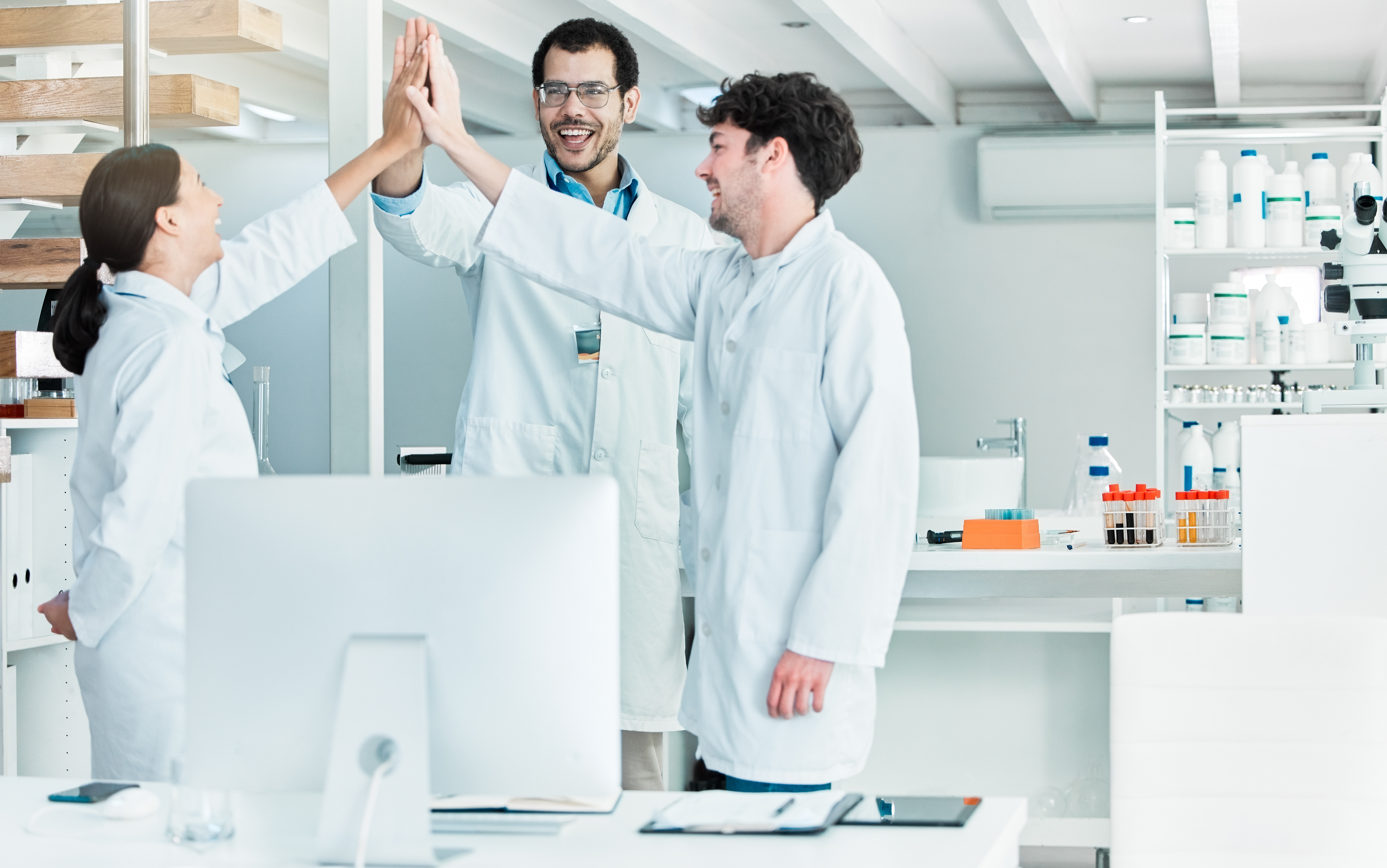  Group of smiling medical professionals in lab coats high-fiving in a modern laboratory setting, symbolizing teamwork and achievement in healthcare.